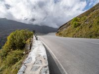 an empty road winds through the mountains below clouds and fogs, with rock walls and vegetation in the foreground