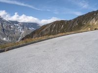 a lone bench is in front of an empty road with mountains in the background with some clouds