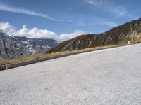 a lone bench is in front of an empty road with mountains in the background with some clouds