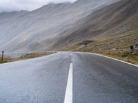 an empty open road leading to mountains with fog above it and a cloudy sky overhead