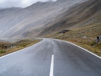 an empty open road leading to mountains with fog above it and a cloudy sky overhead