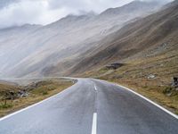 an empty open road leading to mountains with fog above it and a cloudy sky overhead