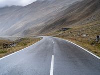 an empty open road leading to mountains with fog above it and a cloudy sky overhead