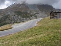 a wooden cabin on the side of a road in front of a mountain range with trees