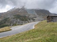 a wooden cabin on the side of a road in front of a mountain range with trees