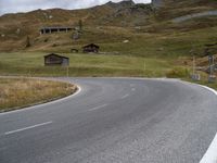 a mountain with an empty road near some houses and a mountain slope behind it that is covered with grass