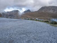 a sheep stands alone on a side walk next to the road with mountains on either side and a cloudy sky overhead