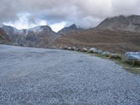 a sheep stands alone on a side walk next to the road with mountains on either side and a cloudy sky overhead