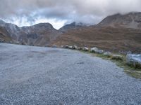 a sheep stands alone on a side walk next to the road with mountains on either side and a cloudy sky overhead