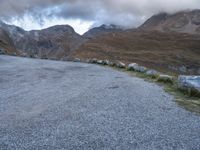 a sheep stands alone on a side walk next to the road with mountains on either side and a cloudy sky overhead