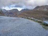 a sheep stands alone on a side walk next to the road with mountains on either side and a cloudy sky overhead