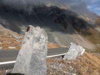 a road surrounded by tall rocks on a hillside with snow capped mountains behind it and a red traffic marker on the side