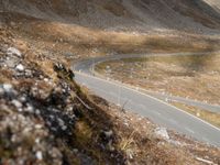 a long narrow mountain road through the grass covered hills above the road is a man riding on a motorcycle