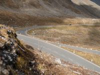 a long narrow mountain road through the grass covered hills above the road is a man riding on a motorcycle