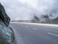 a road with a large rock wall near by it and some fog on top of the mountain