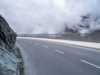 a road with a large rock wall near by it and some fog on top of the mountain