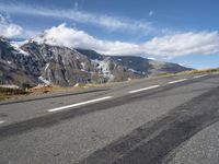 an empty road lined with mountains with snow on the tops of the mountain peaks and white clouds in the sky