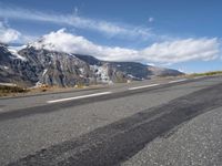 an empty road lined with mountains with snow on the tops of the mountain peaks and white clouds in the sky