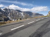 an empty road lined with mountains with snow on the tops of the mountain peaks and white clouds in the sky