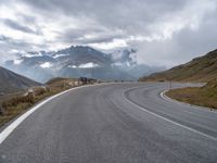 an empty road is winding into the mountains on a cloudy day with clouds flying overhead