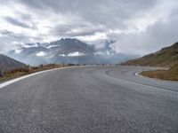 an empty road is winding into the mountains on a cloudy day with clouds flying overhead