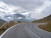 an empty road is winding into the mountains on a cloudy day with clouds flying overhead