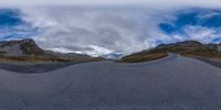 a fish eye view of a long gravel road in mountains surrounding a large cloud,