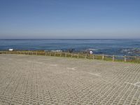 an empty beach and fenced in area with ocean in background, including a person on a bench at the end