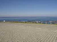 an empty beach and fenced in area with ocean in background, including a person on a bench at the end