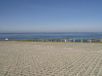 an empty beach and fenced in area with ocean in background, including a person on a bench at the end