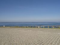 an empty beach and fenced in area with ocean in background, including a person on a bench at the end