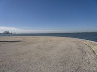 a lone beach sits near the ocean with a bench at one end of it that's facing a city skyline