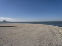 a lone beach sits near the ocean with a bench at one end of it that's facing a city skyline