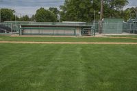 there is a baseball field with several dug outs behind the baseball field in which there is a bench, green grass, and the stadium has a sky