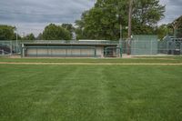 there is a baseball field with several dug outs behind the baseball field in which there is a bench, green grass, and the stadium has a sky