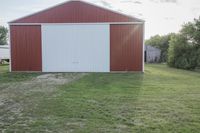 an older barn with a green grass lawn in the foreground and large red building behind it