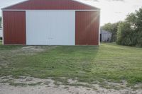 an older barn with a green grass lawn in the foreground and large red building behind it