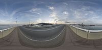 a fish eye lens captures the highway next to the sea and mountains and in the distance the sky is cloudy