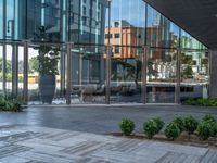 a glass walled building is reflecting the surrounding buildings and trees in the courtyard area with tables and chairs