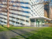 a building with a glass walkway leading up to it surrounded by tall buildings, a grassy lawn, and trees on one side of the building