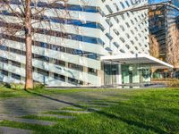 a building with a glass walkway leading up to it surrounded by tall buildings, a grassy lawn, and trees on one side of the building