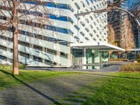 a building with a glass walkway leading up to it surrounded by tall buildings, a grassy lawn, and trees on one side of the building