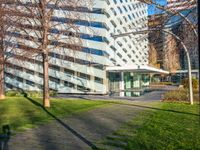 a building with a glass walkway leading up to it surrounded by tall buildings, a grassy lawn, and trees on one side of the building