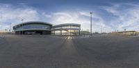 a curved building in a parking lot under a cloudy blue sky, with a glassy roof