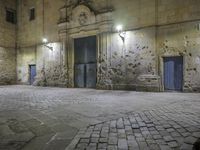 a stone walkway in front of an empty building at night with blue doorways and brick walling