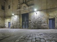 a stone walkway in front of an empty building at night with blue doorways and brick walling