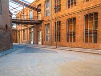 a lone empty street in an old brick building complex with a metal walkway between the buildings