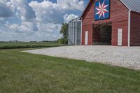 a barn on top of a gravel road next to grass and trees in front of a blue sky