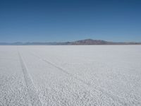 a barren plain with tire tracks in the middle of it is surrounded by mountains and a clear blue sky