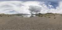 large cloud over water and sand with mountain in distance behind it and field in the foreground